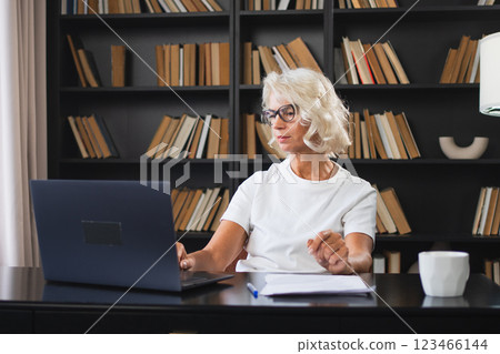 Confident stylish middle aged senior woman using laptop at workplace. Older mature lady businesswoman sitting at office table with laptop and paper document. Boss leader teacher professional worker 123466144
