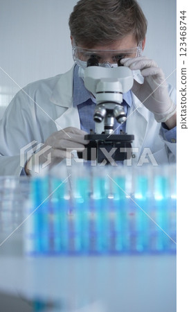 Male scientist researcher wearing lab coat, white protective gloves, medical mask and glasses, is using microscope in modern laboratory setup, portrait view. Low key lighting photo. Science Male scientist researcher wearing lab coat, white protective gloves, medical mask and glasses, is using microscope in modern laboratory setup, portrait view. Low key lighting photo. Science 123468744