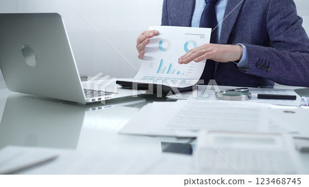 Businessman wearing blue suit is working with laptop while examining financial data, using charts and graphs to make informed decisions in a professional office setting. Low lighting, close-up view Businessman wearing blue suit is working with laptop while examining financial data, using charts and graphs to make informed decisions in a professional office setting. Low lighting, close-up view 123468745