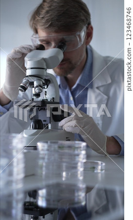Man scientist wearing lab coat, white gloves and protective glasses using microscope doing research in modern laboratory, portrait view. Low key lighting photo. Science and Medicine Man scientist wearing lab coat, white gloves and protective glasses using microscope doing research in modern laboratory, portrait view. Low key lighting photo. Science and Medicine 123468746