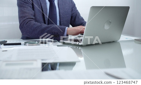 Businessman wearing blue suit is working with laptop, calculator and documents at the glass table in office set. Low lighting, close-up view. Business people and audit concept Businessman wearing blue suit is working with laptop, calculator and documents at the glass table in office set. Low lighting, close-up view. Business people and audit concept 123468747