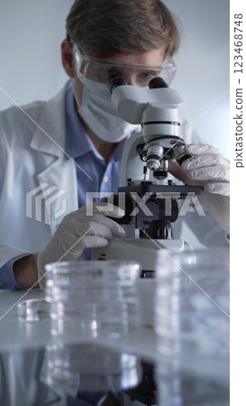 Man scientist wearing lab coat, white gloves, medical mask and protective glasses using microscope doing research in modern laboratory, portrait view. Low key lighting photo. Science and Medicine Man scientist wearing lab coat, white gloves, medical mask and protective glasses using microscope doing research in modern laboratory, portrait view. Low key lighting photo. Science and Medicine 123468748