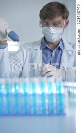 Male scientist researcher, wearing lab coat, white medical gloves, mask and protective glasses is using micropipette filling test tubes in modern laboratory. Low key lighting. Science concept Male scientist researcher, wearing lab coat, white medical gloves, mask and protective glasses is using micropipette filling test tubes in modern laboratory. Low key lighting. Science concept 123468749