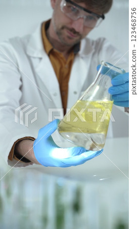 Man scientist, wearing blue gloves and protective glasses, is holding a yellow chemical solution inside Erlenmeyer flask in a laboratory, vertical portrait view. Science and medicine 123468756