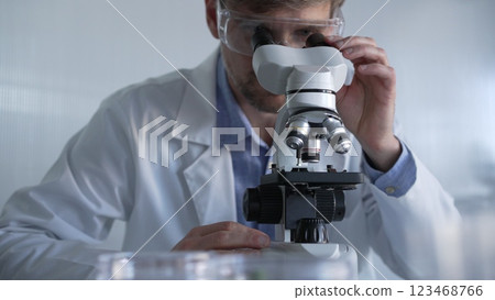 Male scientist wearing lab coat and protective glasses using microscope doing research in modern laboratory setup, portrait view. Low key lighting photo. Science and Medicine concept Male scientist wearing lab coat and protective glasses using microscope doing research in modern laboratory setup, portrait view. Low key lighting photo. Science and Medicine concept 123468766