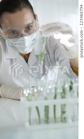 Woman scientist wearing a lab coat, white gloves, face mask and protective glasses, is holding a test tube with plants inside, vertical portrait view. Science and medicine 123468794
