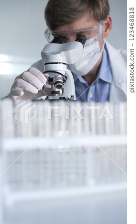 Man scientist wearing lab coat, white gloves, medical mask and protective glasses using microscope doing research in modern laboratory, portrait view. Low key lighting photo. Science and Medicine 123468818