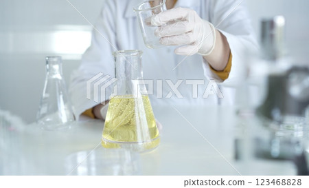 A scientist, wearing a lab coat and white protective gloves, is pouring a yellow oily liquid from one beaker to another in laboratory, close up. Medicine and science concept 123468828
