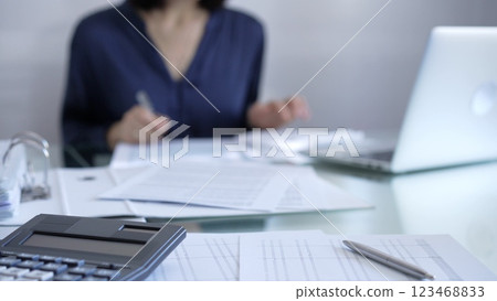 Black calculator, pen and documents are lying on accountant's desk. Businesswoman wearing blue dress is working with documents on the background. Business and audit concept 123468833