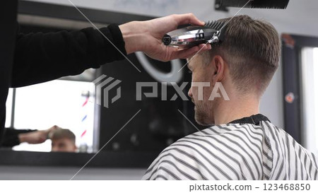 Professional barber trimming male client's hair using electric razor, creating precise haircut in contemporary barbershop setting with sleek styling tools Professional barber trimming male client's hair using electric razor, creating precise haircut in contemporary barbershop setting with sleek styling tools 123468850