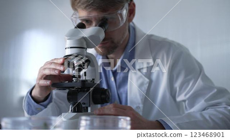 Male scientist wearing lab coat and protective glasses using microscope doing research in modern laboratory setup, portrait view. Low key lighting photo. Science and Medicine concept 123468901