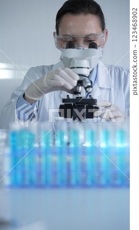 Woman scientist wearing lab coat, white protective gloves and glasses, is using microscope in modern laboratory setup, portrait view. Low key lighting photo. Science and Medicine concept 123468902