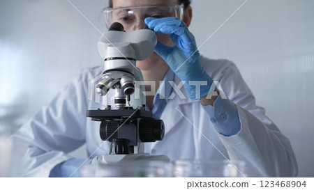 Woman scientist wearing lab coat, white protective gloves and glasses, is using microscope in modern laboratory setup, portrait view. Low key lighting photo. Science and Medicine 123468904
