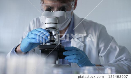 Male scientist researcher wearing blue gloves, protective glasses and medical mask, is using microscope in modern laboratory, portrait view. Low key lighting photo. Science and Medicine Male scientist researcher wearing blue gloves, protective glasses and medical mask, is using microscope in modern laboratory, portrait view. Low key lighting photo. Science and Medicine 123468907