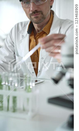 Man science researcher, wearing a lab coat and protective glasses, drips liquid from a pipette into a test tube with a green plant. His face is focused, captured in a vertical portrait. Science and 123468909