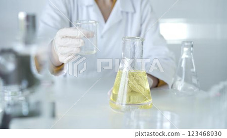 A scientist, wearing a lab coat and white protective gloves, is pouring a yellow oily liquid from one beaker to another in laboratory, close up. Medicine and science concept A scientist, wearing a lab coat and white protective gloves, is pouring a yellow oily liquid from one beaker to another in laboratory, close up. Medicine and science concept 123468930