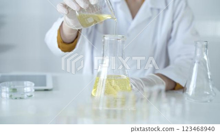 A scientist, wearing a lab coat and white protective gloves, is pouring a yellow oily liquid from one beaker to another in laboratory, close up. Medicine and science concept A scientist, wearing a lab coat and white protective gloves, is pouring a yellow oily liquid from one beaker to another in laboratory, close up. Medicine and science concept 123468940