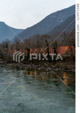 River Lodges Mountains Dusk: Illuminated riverside lodges nestled in mountainous terrain at twilight, offering tranquil getaway. 123471388