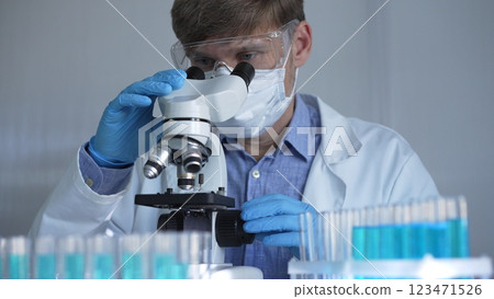 A scientist man, wearing a lab coat, mask, blue gloves and safety glasses, is using a binocular microscope surrounded by test tubes in laboratory. Portrait view. Medicine and science A scientist man, wearing a lab coat, mask, blue gloves and safety glasses, is using a binocular microscope surrounded by test tubes in laboratory. Portrait view. Medicine and science 123471526