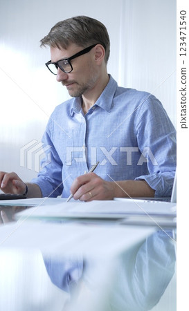 Businessman or accountant, wearing blue casual shirt and glasses, is working on tax calculations, note taking, and financial reports in the office. Audit and taxes in business 123471540