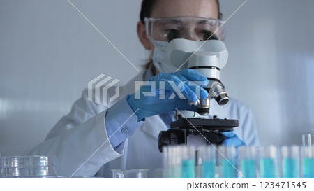 Scientist woman, wearing a lab coat, mask, blue gloves and safety glasses, is adjusting a microscope surrounded by test tubes in laboratory. Medicine and science concept Scientist woman, wearing a lab coat, mask, blue gloves and safety glasses, is adjusting a microscope surrounded by test tubes in laboratory. Medicine and science concept 123471545
