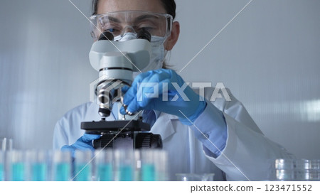 Scientist woman, wearing a lab coat, mask, blue gloves and safety glasses, is adjusting a microscope surrounded by test tubes in laboratory. Medicine and science concept 123471552
