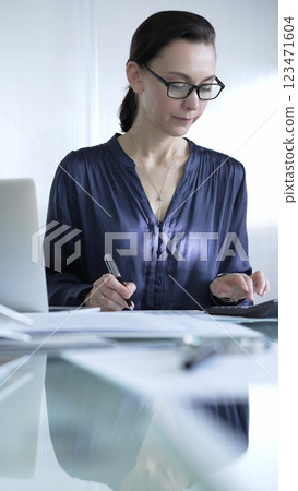 Professional woman accountant in glasses using a calculator and taking notes at a glass desk in the office. Audit and taxes in business 123471604
