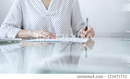Businesswoman in white dotted blouse is signing a contract at a sleek glass desk in a modern office, showcasing professionalism and focus in her work environment. Business people concept Businesswoman in white dotted blouse is signing a contract at a sleek glass desk in a modern office, showcasing professionalism and focus in her work environment. Business people concept 123471731