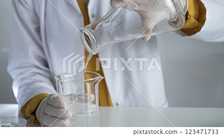 Unknown scientist wearing a lab coat and gloves is carefully pouring a liquid from one beaker into another, close up. Science and medicine concepts 123471733