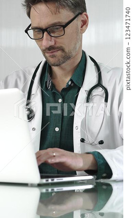 A male doctor in a green shirt under a medical coat is using technology in healthcare. Man is typing on a laptop on a glass desk in the clinic, vertical portrait view. Medicine A male doctor in a green shirt under a medical coat is using technology in healthcare. Man is typing on a laptop on a glass desk in the clinic, vertical portrait view. Medicine 123471740