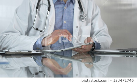 Doctor man, wearing a blue shirt under a medical white coat, is using digital tablet in modern healthcare setting, interacting with patient data, close-up. Medicine and health care 123471746