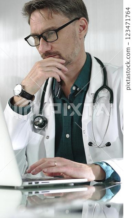 A male doctor in a green shirt under a medical coat is thinking while using technology in healthcare. Man is typing on a laptop on a glass desk in the clinic, vertical portrait view. Medicine concept 123471764