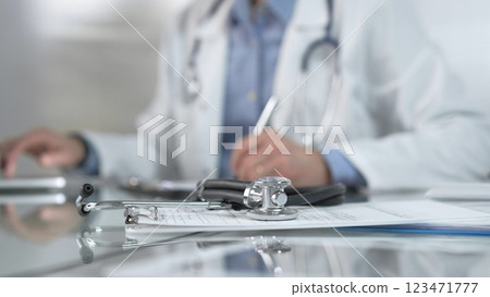 Stethoscope and medication history records form are on a glass table before a male doctor working with laptop in clinical office background. Medicine concept 123471777