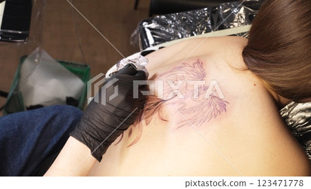 Professional tattoo artist wearing black gloves drawing intricate design on woman's back while working in sleek, well lit tattoo studio with modern equipment 123471778