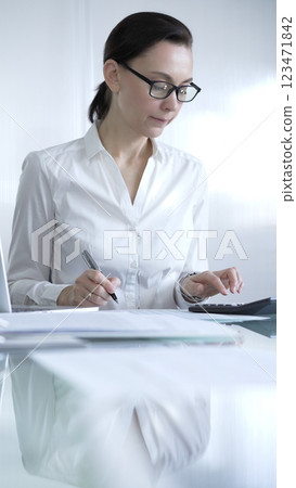 Businesswoman or accountant, wearing white casual blouse and glasses, is working on tax calculations, note taking, and financial reports in the office. Audit and taxes in business 123471842