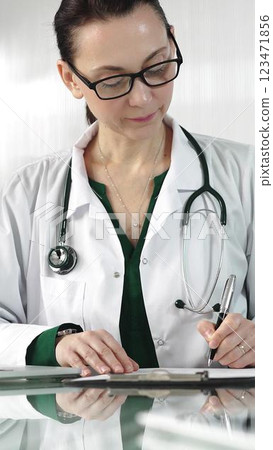 Female doctor in a green blouse under a medical coat is filling up medication history records on a glass desk in the clinic, vertical portrait view. Medicine concept Female doctor in a green blouse under a medical coat is filling up medication history records on a glass desk in the clinic, vertical portrait view. Medicine concept 123471856