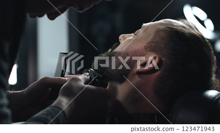 Skilled barber grooming client's beard using electric razor, delivering precise styling within contemporary barbershop setting Skilled barber grooming client's beard using electric razor, delivering precise styling within contemporary barbershop setting 123471943