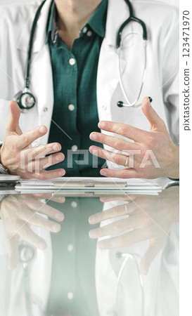 Doctor man wearing a stethoscope and lab coat is gesturing with hands while explaining a diagnosis to a patient across a glass desk with a laptop. Medicine concept 123471970