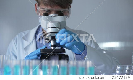 A scientist man, wearing a lab coat, mask, blue gloves and safety glasses, is using a binocular microscope surrounded by test tubes in laboratory. Portrait view. Medicine and science 123472139