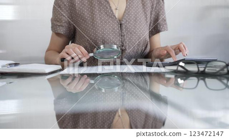 Businesswoman accountant in brawn dotted blouse reviewing financial reports with magnifying glass and calculator, conducting audit and analyzing data in office. Taxes, audit in business 123472147