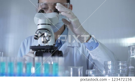 A scientist woman, wearing a lab coat, mask, white gloves and safety glasses, is adjusting a microscope surrounded by test tubes in laboratory A scientist woman, wearing a lab coat, mask, white gloves and safety glasses, is adjusting a microscope surrounded by test tubes in laboratory 123472156