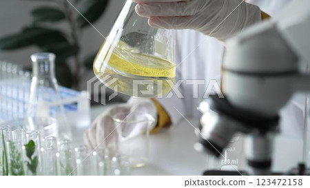Unknown scientist wearing white protective gloves is carefully shaking up a yellow chemical solution inside an Erlenmeyer flask in a laboratory, close up. Science and medicine 123472158