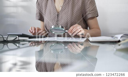 Businesswoman accountant in brawn dotted blouse reviewing financial reports with magnifying glass and calculator, conducting audit and analyzing data in office. Taxes, audit in business 123472266