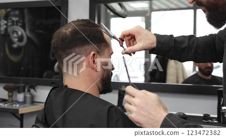 Skilled barber trimming client's hair using professional scissors, creating contemporary hairstyle in modern barbershop setting with precise, professional technique. 123472382