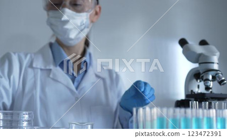 Scientist woman, wearing a lab coat, mask, blue gloves and safety glasses, is using a micropipette to drop a liquid into a test tubes. Medicine and science Scientist woman, wearing a lab coat, mask, blue gloves and safety glasses, is using a micropipette to drop a liquid into a test tubes. Medicine and science 123472391