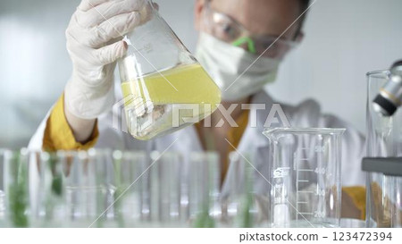Woman scientist researcher wearing white protective gloves is shaking up a yellow chemical solution inside an Erlenmeyer flask in a laboratory setting, portrait view. Science and medicine 123472394