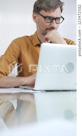 Focused businessman in yellow casual shirt is using laptop and thinking with hand on chin, working on a project in a modern office, vertical portrait. Business people concept 123472582