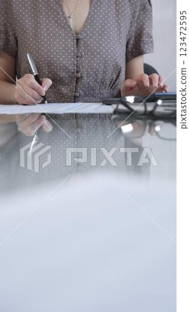 Businesswoman accountant, wearing brawn dotted blouse, is using a calculator at sleek glass desk in a contemporary office, vertical view. Taxes, audit in business 123472595
