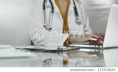 Female doctor, wearing yellow blouse under a medical coat, is using technology in healthcare, close up of hands typing on laptop with stethoscope, glasses, and clipboard on desk. Medicine concept 123472624