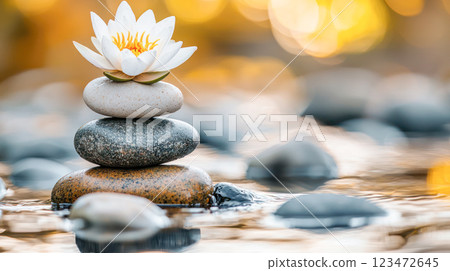 Stacked spa stones with a delicate white lotus flower floating on a calm water surface, surrounded by smooth pebbles and illuminated by a warm golden bokeh background, symbolizing peace and balance. 123472645